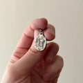 A hand holding a small, irregularly shaped silver pendant engraved with the letter 'E', shown against a plain, light beige background.